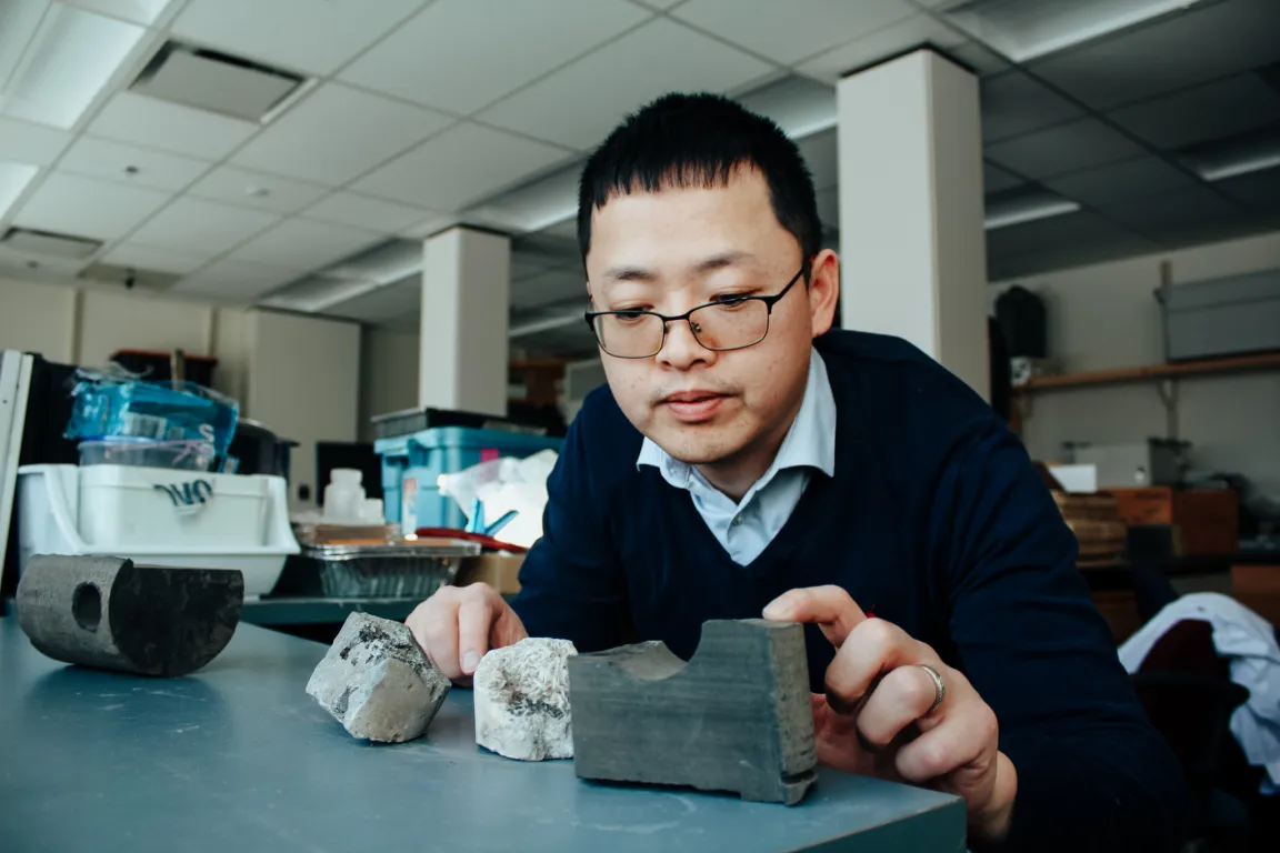 Researchers with glasses wearing blue shirt and navy sweater leans over desk looking at rock core sample.