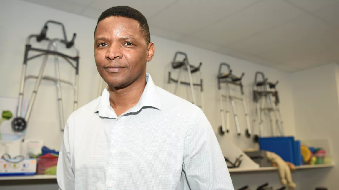 Taru Manyanga stands in front of physical therapy equipment hanging on an ivory wall.