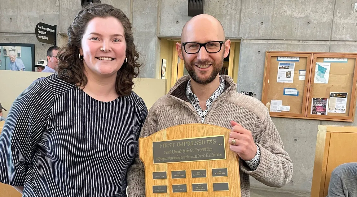 Two people stand together in an atrium with the person on the right holding an award plaque.