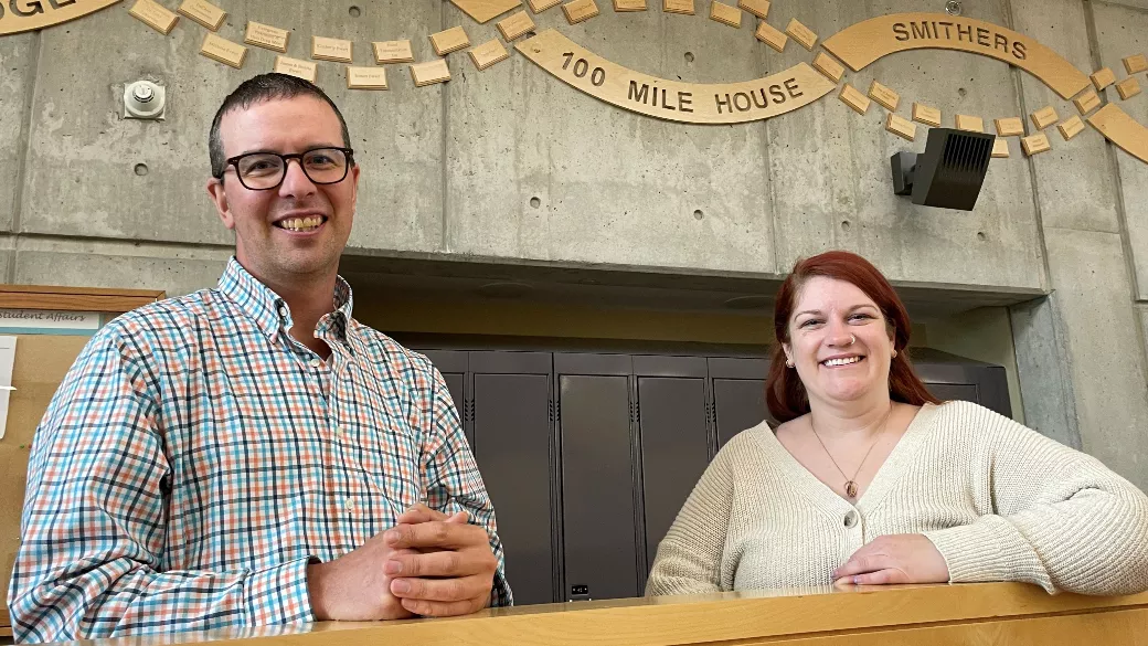 Two people stand in front of a background that features a wood DNA design on the wall.