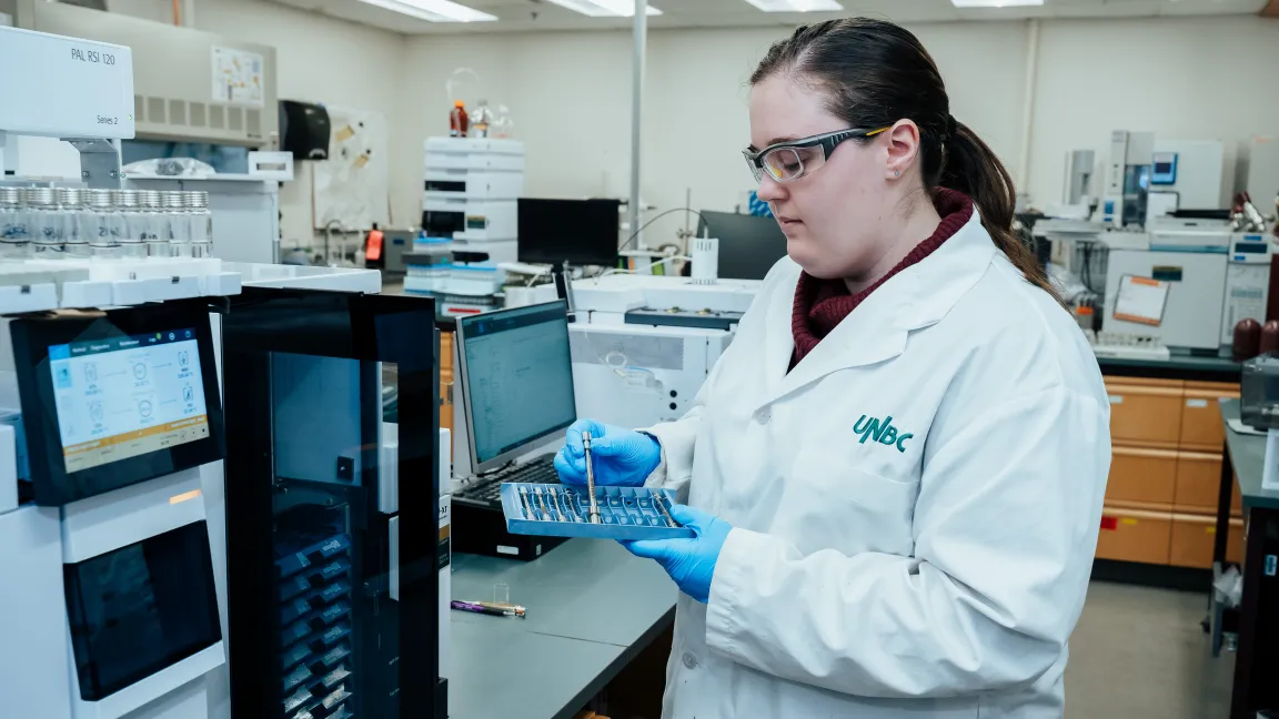 Mya Schouwenburg, an Analytical Lab Specialist at UNBC's Northern Analytical Lab Services (NALS), wearing a lab coat and safety glasses, prepares samples for analysis using laboratory equipment. The lab workspace is filled with scientific instruments and computer monitors.