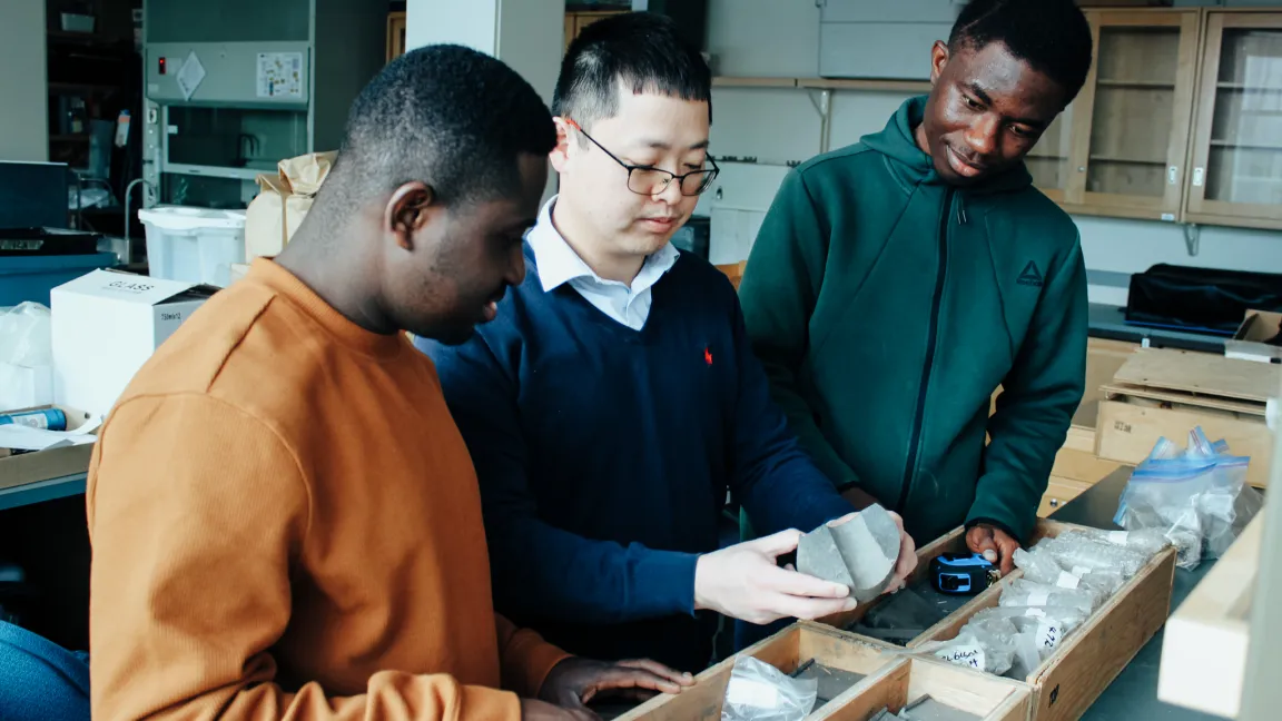 Three people stand at a table in a laboratory setting looking at rock core samples.