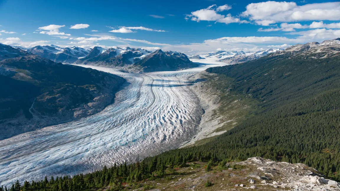 Aerial view showing glacier in a mountain valley. Snow-capped mountain peaks with blue sky and clouds in background.