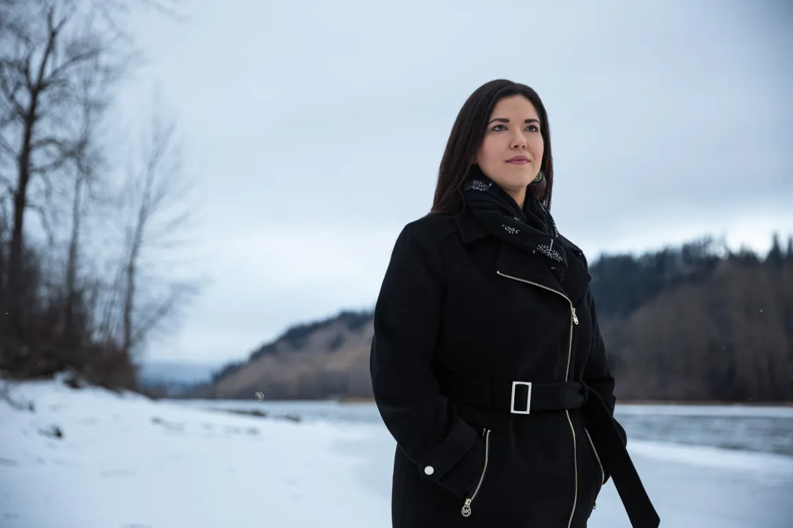 Dr. Terri Aldred stands on a snow-covered riverbank, with forested hills and overcast skies in the background.