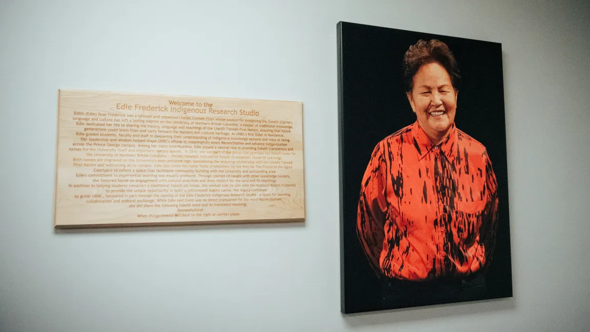 Photo shows a portrait and plaque on a wall. The portrait features a person in a red and black top and black pants on a black canvas. The wooden plaque on the left side contains an engraved description.