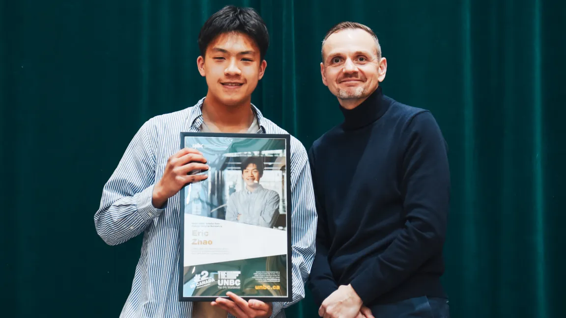 Two people standing in front of dark green curtain. One holds framed poster.
