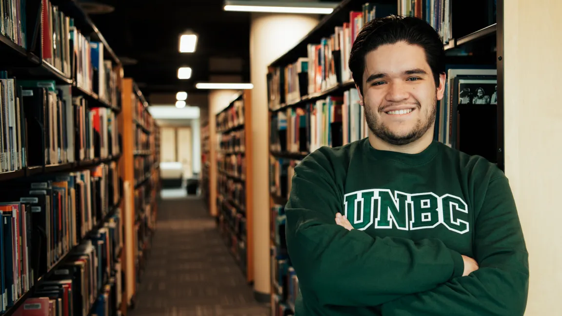 Student wearing green UNBC crewneck sweatshirt leans with arms folded against library shelving with books in background