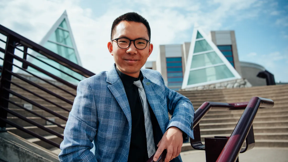 Person wearing black dress shirt and blue striped blazer poses on stairs with skylights in background.