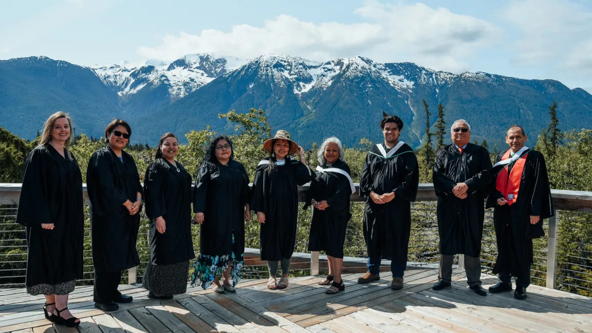 Nine people standing in black academic robes on an outdoor platform with mountains in background.