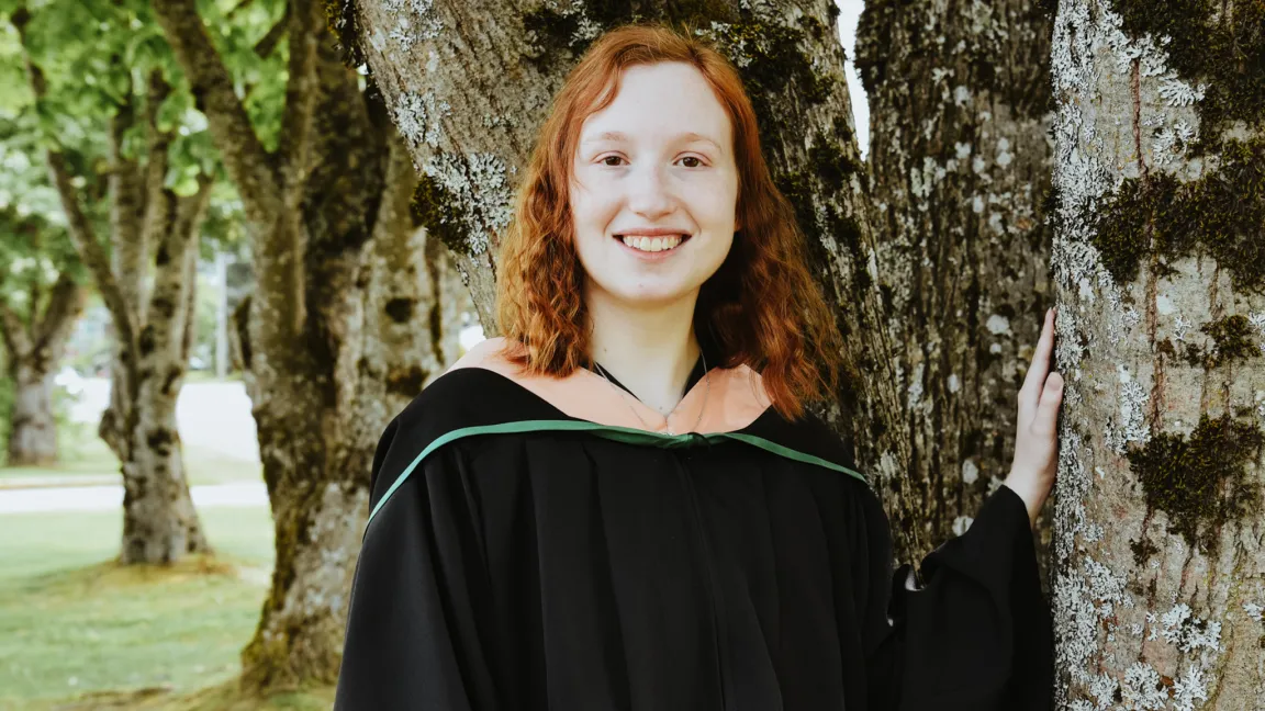 Student wearing black academic robe and hood with peach-coloured stripe stands outside with hand on a tree trunk. Trees in background
