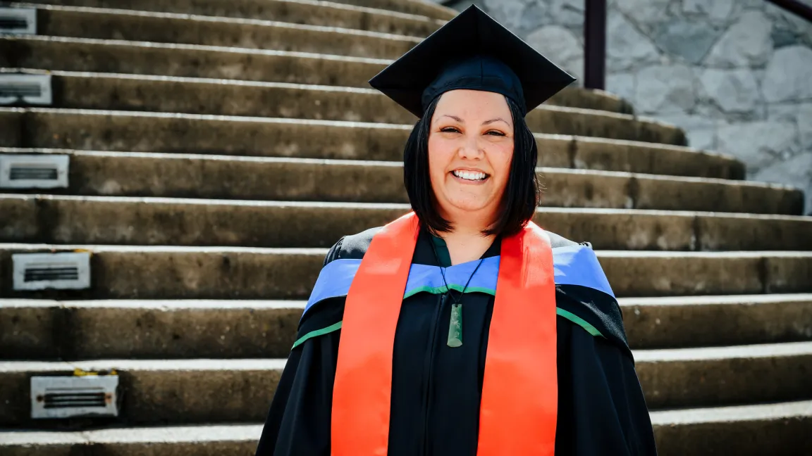 Laura Mueller smiles in her graduation regalia in front of a stone staircase on UNBC’s Prince George campus.