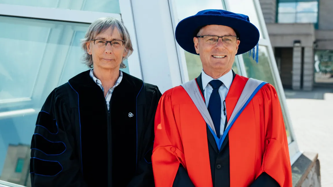 Dr. Kathy Lewis and Dr. Kerry Reimer stand in academic regalia outside a UNBC building during the Professor Emeritus recognition event.