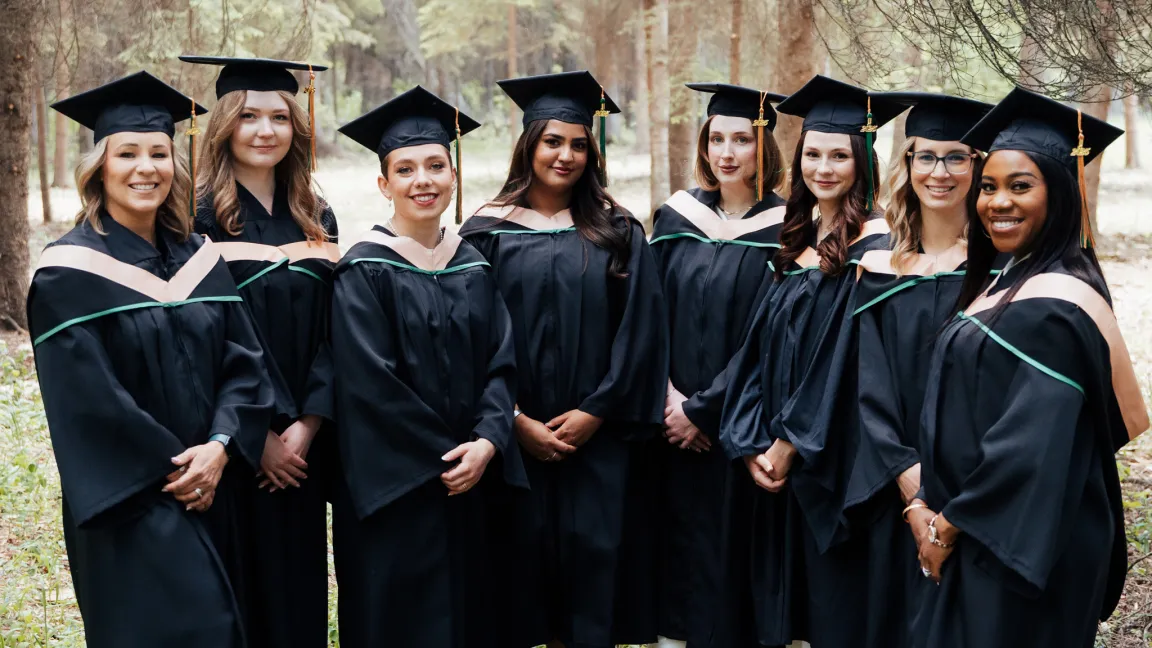 Eight graduates from UNBC’s Northern Baccalaureate Nursing Program stand together in convocation regalia among trees near the Fort St. John campus.