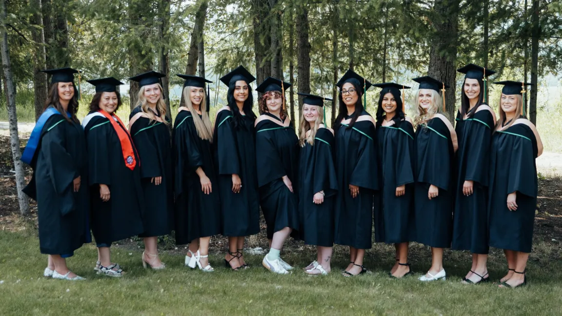 A group of graduates wearing black gowns and mortarboards stand side by side outdoors in front of a backdrop of tall trees. They are smiling and dressed for a graduation celebration, with some wearing hoods or stoles in various colours.