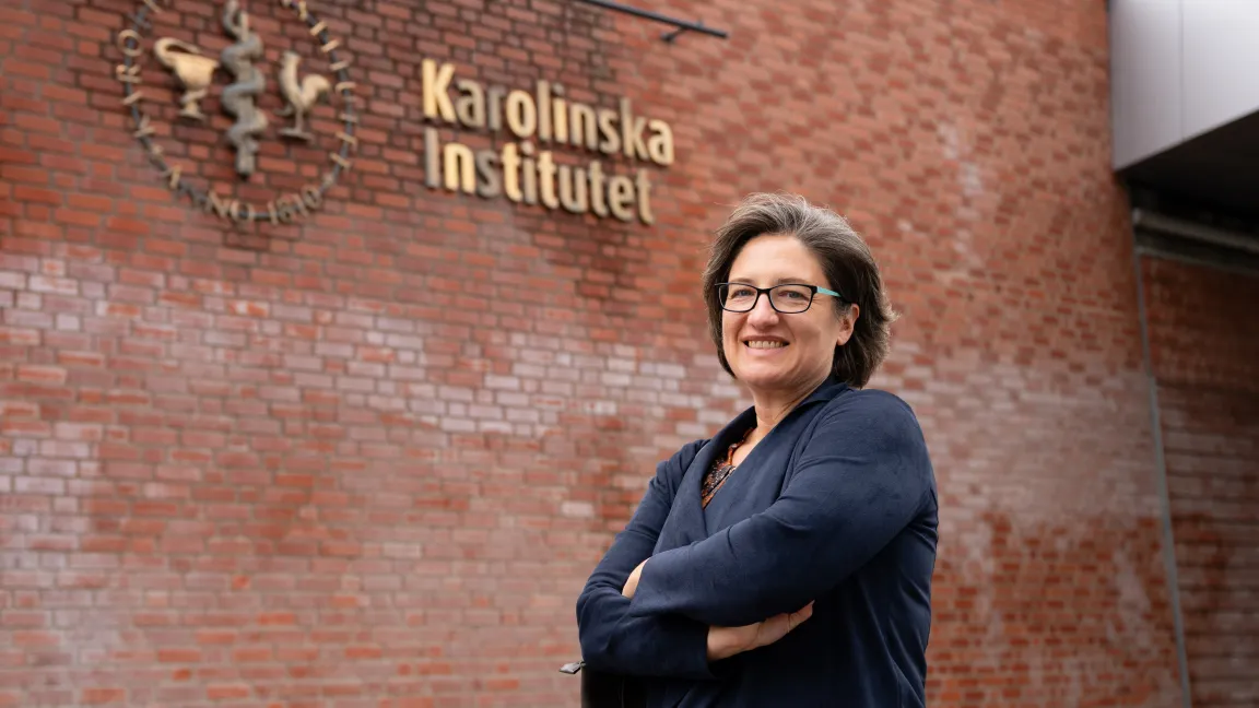 Andrea Gingerich, dressed in a navy cardigan, stands in front of a fading red brick wall with the words Karolinska Institutet on it.