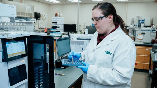 Mya Schouwenburg, an Analytical Lab Specialist at UNBC's Northern Analytical Lab Services (NALS), wearing a lab coat and safety glasses, prepares samples for analysis using laboratory equipment. The lab workspace is filled with scientific instruments and computer monitors.
