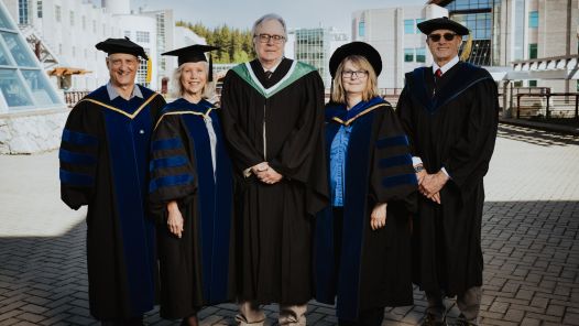 Five people standing in a row wearing black academic regalia with hoods