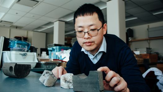 Researchers with glasses wearing blue shirt and navy sweater leans over desk looking at rock core sample.