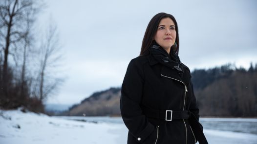 Dr. Terri Aldred stands on a snow-covered riverbank, with forested hills and overcast skies in the background.