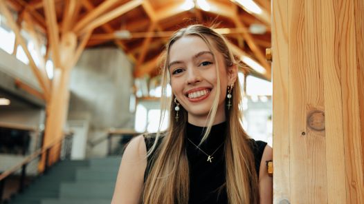 Person wearing black sleeveless top, gold necklace with letter K and earrings leans against a wooden column with stairs and wooden-beamed ceiling in background.