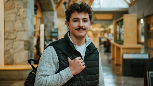 Person wearing a grey shirt and navy vest holds black backpack strap near their shoulder and poses for photo in a hallway.