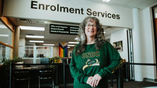 A person wearing a UNBC Indigenous logo shirt standing in front of the Enrolment Services office. 
