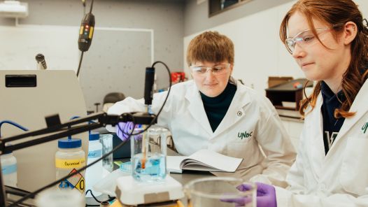 Two students in lab coats, wearing safety glasses work in a lab at one of Canada's top undergraduate research universities.