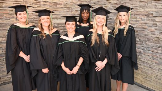 Six people wearing academic regalia pose for photo in front of rock wall.