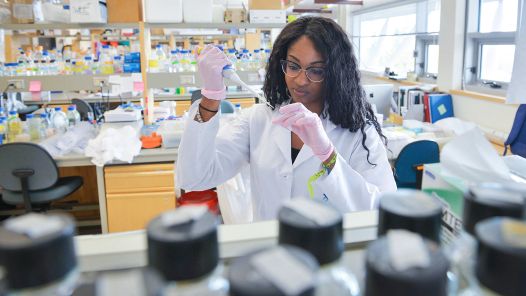 Researcher wearing white lab coat in a laboratory setting wearing gloves and working with a syringe and test tube.