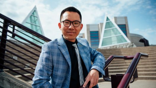 Person wearing black dress shirt and blue striped blazer poses on stairs with skylights in background.