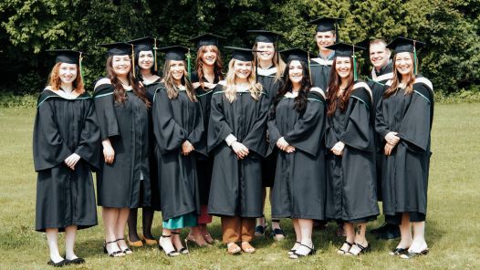 Large group of graduate students wearing black academic robes and mortarboards stands outdoors.