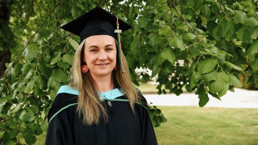 A woman in graduation robes and mortarboard stands in front of green leafy trees, smiling.