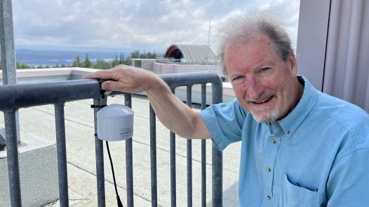 A person on a roof of a building, holding a railing. Attached to the railing is an air quality monitor 