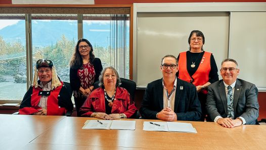 Six people around a table, some in traditional Indigenous regalia at a document signing ceremony