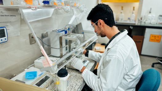 A person working in a lab, wearing a lab coat and safety glasses. 