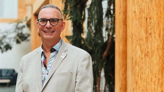 A person standing in the atrium of a well lit building, wearing a suit jacket with a UNBC pin