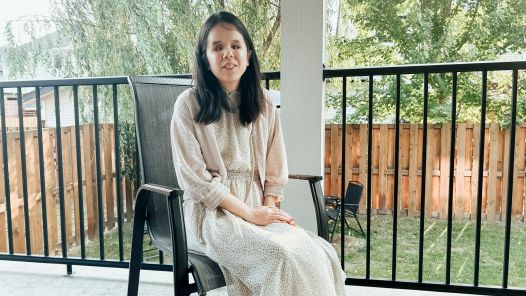 Kaho Michihara sits on a patio chair in a light-patterned dress and textured cardigan, with a wooden fence and trees in the background.