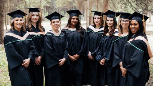 Eight graduates from UNBC’s Northern Baccalaureate Nursing Program stand together in convocation regalia among trees near the Fort St. John campus.