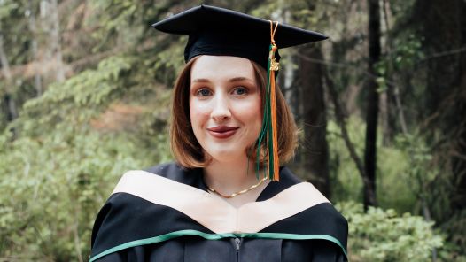 Megan Wilson wears her graduation gown and cap with a green and gold tassel while standing in a forested area. She is looking at the camera and smiling softly.