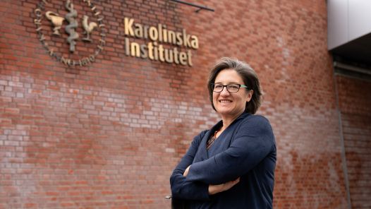 Andrea Gingerich, dressed in a navy cardigan, stands in front of a fading red brick wall with the words Karolinska Institutet on it.