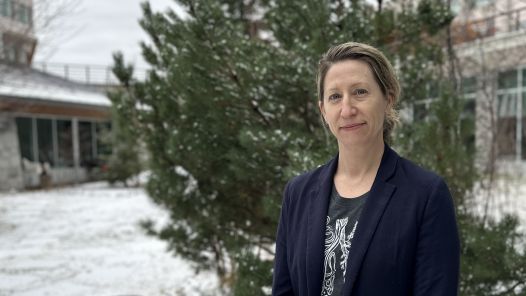 Person wearing grey shirt with white logo and blue blazer stands outside with trees, snow and building in background.