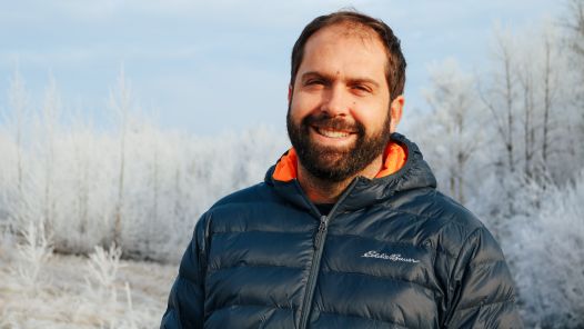 Person wearing navy jacket with orange-lined hood stands outside with snow-covered trees in background.