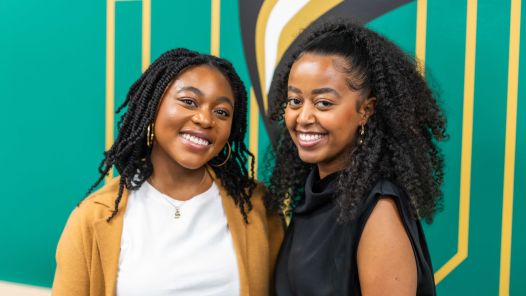 Portrait photo of two people standing in front of a green wall with silver lettering. The photo is too close to the wall to make out the letters/ 