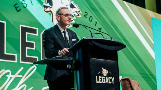 UNBC President Dr. Geoff Payne speaks at the podium during Timberwolves Legacy Night, standing in front of a green backdrop featuring the event logo and the word “LEGACY.”
