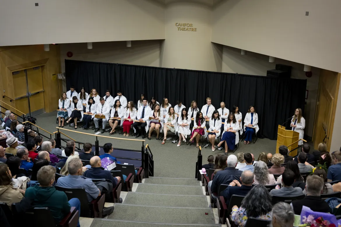 The NMP Class of 2025 sits in two rows of chairs wearing their white coats in front of a populated auditorium venue.