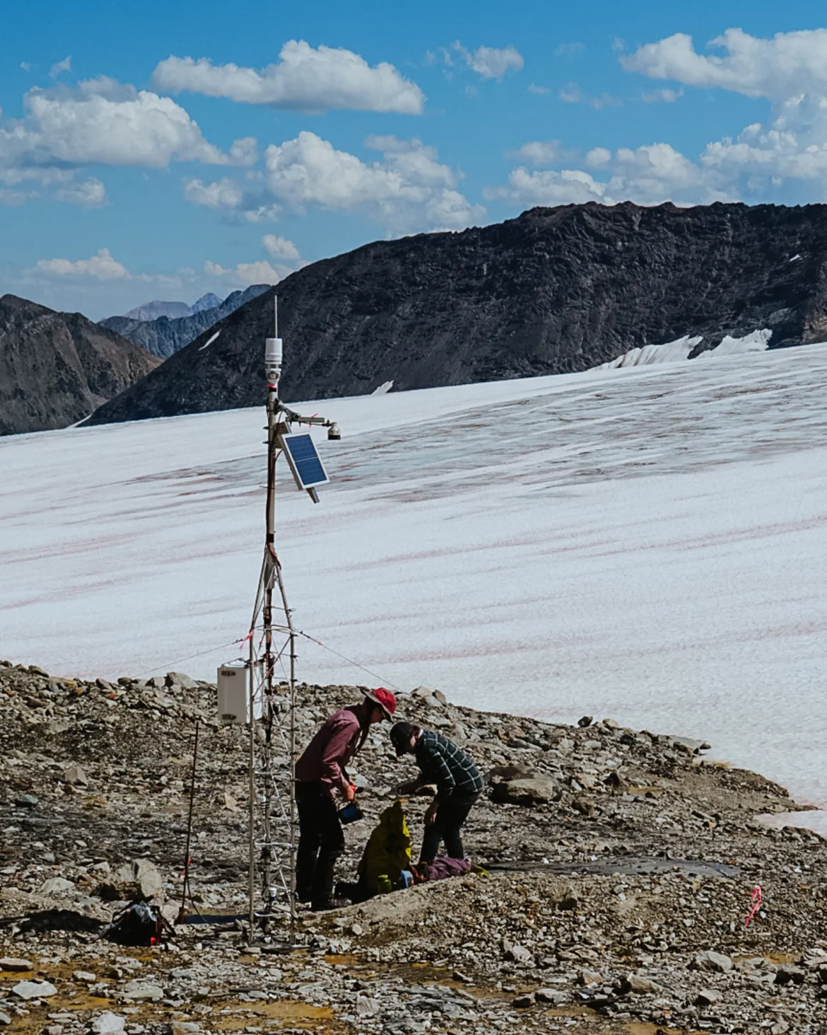Two student researchers kneeling beside a snow monitoring station. Mountains in background.