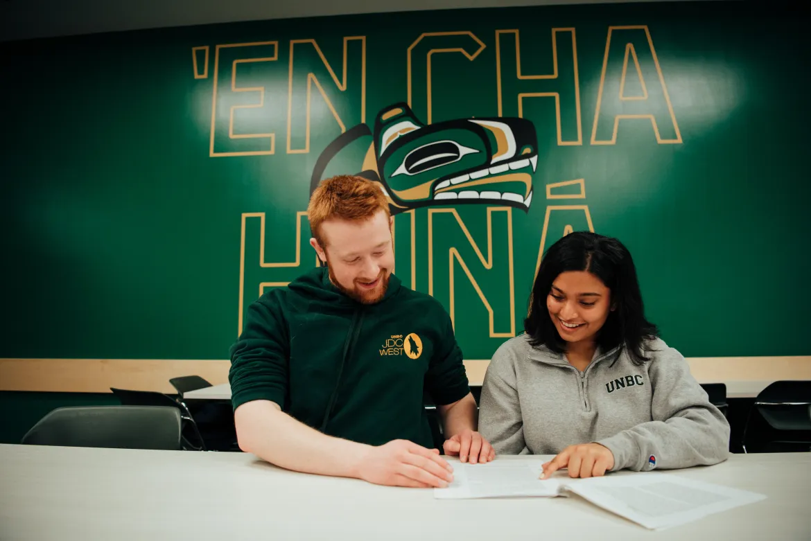 Two students with UNBC Logo in the back, male student with red hair and female student with darker skin and black hair