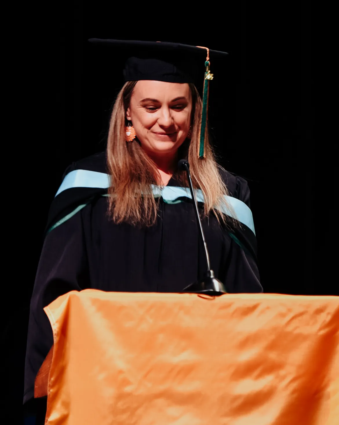 Person standing at podium in black academic regalia with blue stripe on hood.