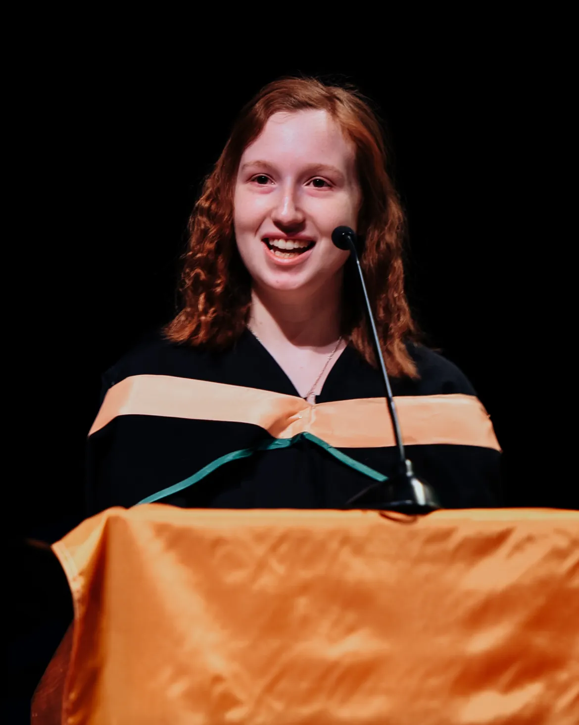 Person standing at a podium wearing black academic robe with orange stripe on hood.