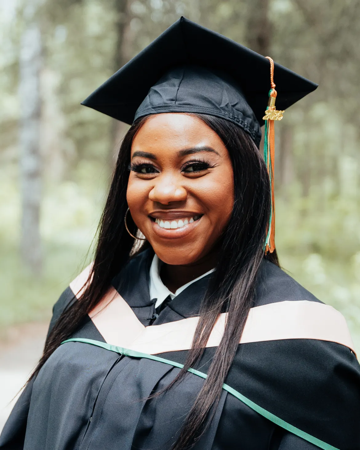 Blessing Chioma Nwanekezie smiles in her graduation regalia during UNBC’s Northeast Campus Convocation, with trees softly blurred in the background.